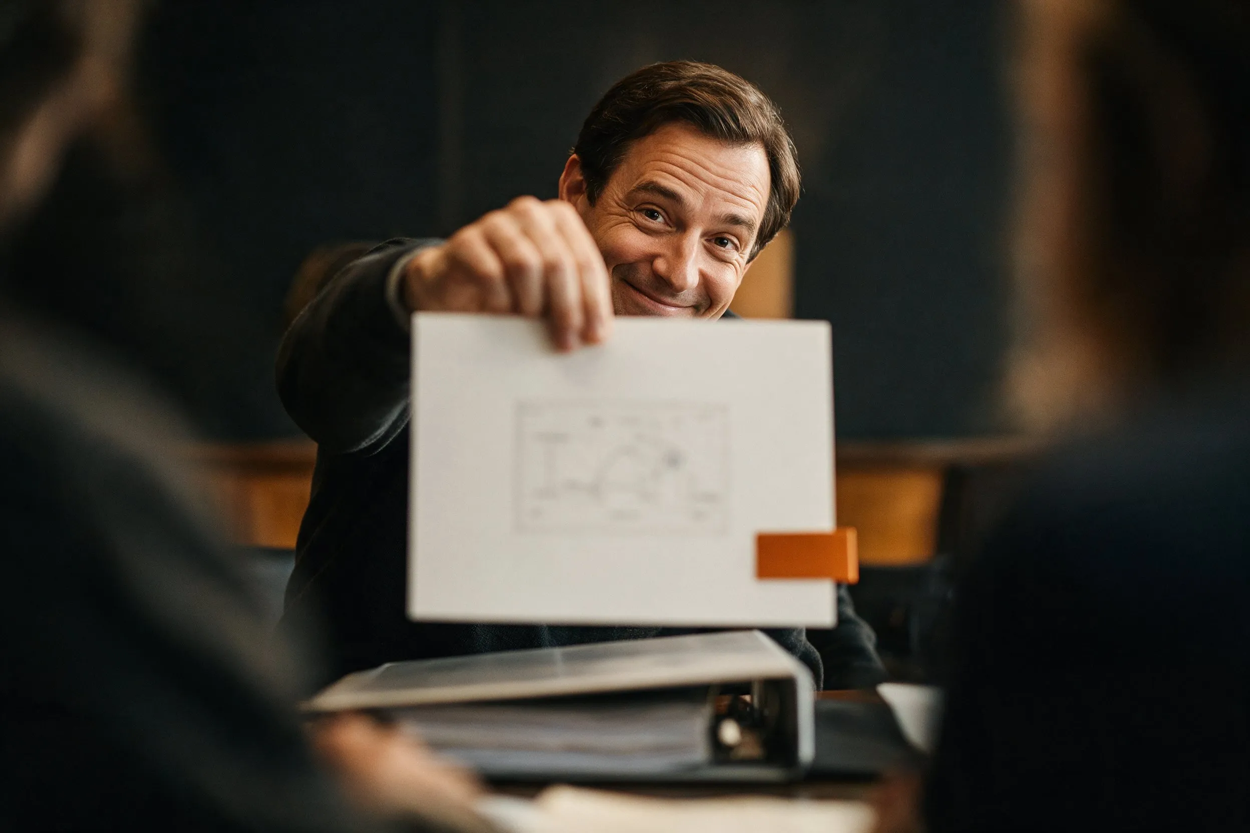 A presenter smiles over the edge of a single printed card held toward the camera, a thick binder closed on the table below.