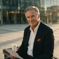 A silver-haired executive on a plaza bench at golden hour meets the camera with a calm, knowing half-smile, coffee beside him.